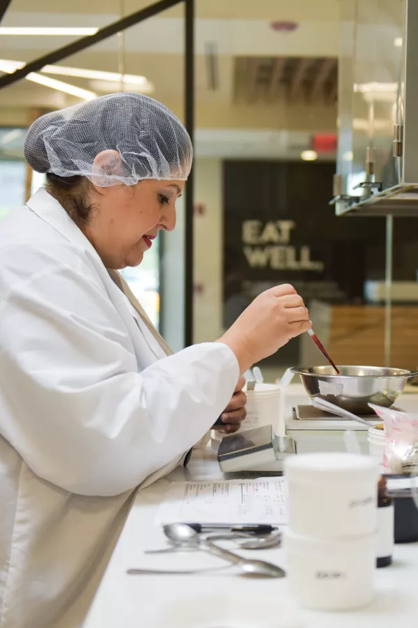 A person in a lab coat and hairnet stirs a mixture in a metal bowl on a counter with containers, utensils, and a clipboard nearby.
