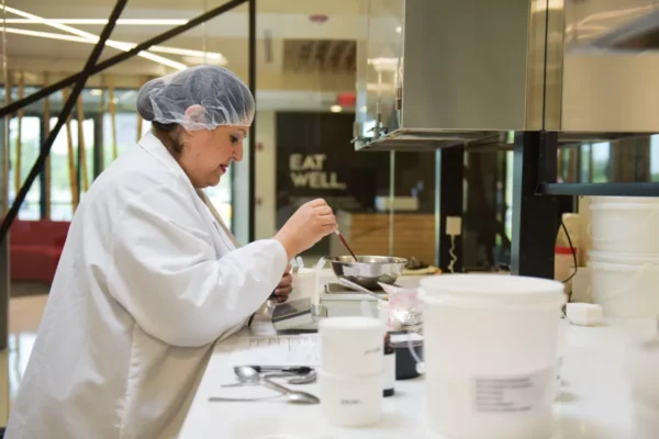 A person in a lab coat and hairnet uses a spoon to mix ingredients in a metal bowl at a laboratory counter with various containers and equipment.