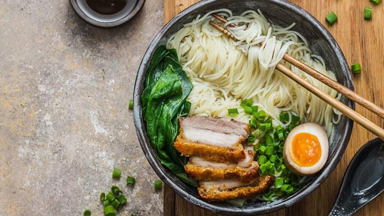 A bowl of ramen noodles with sliced pork, spinach, green onions, and half a soft-boiled egg, served with chopsticks and a spoon on a wooden board.