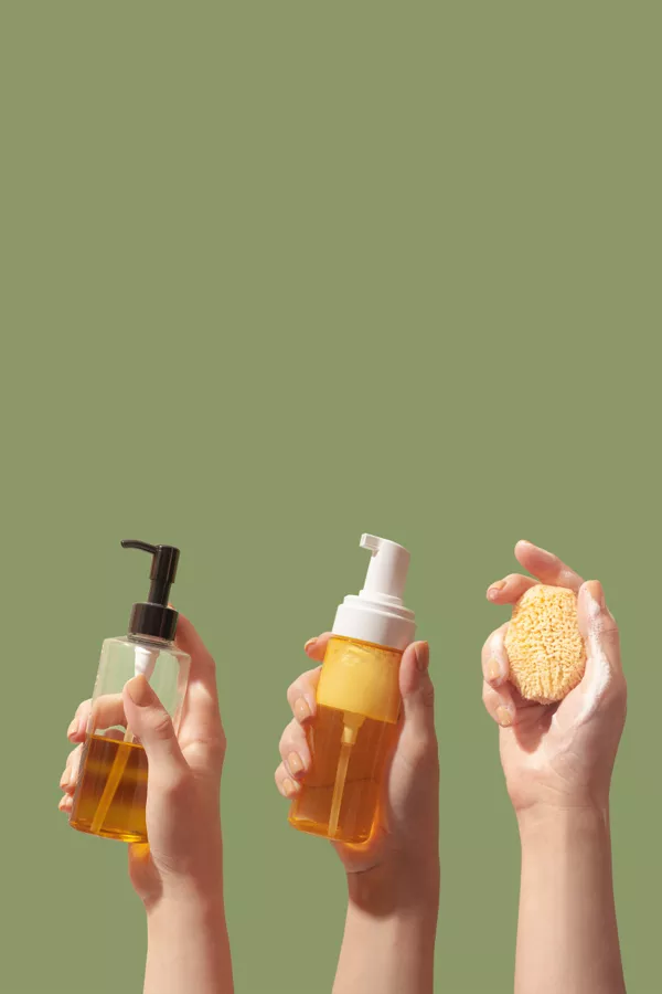 Three hands hold skincare items: a pump bottle, a foaming dispenser, and a natural sponge, all containing or covered in orange liquid, against a plain green background.