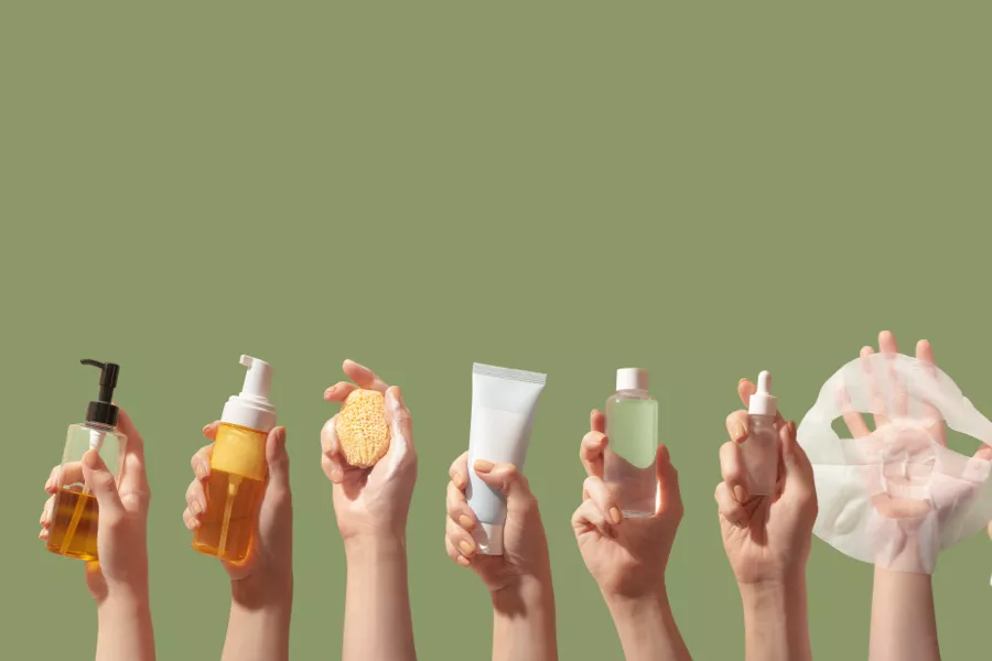 Six hands each hold a different skincare product: liquid soap, foam cleanser, sponge, cream tube, toner bottle, and a sheet mask against a plain green background.