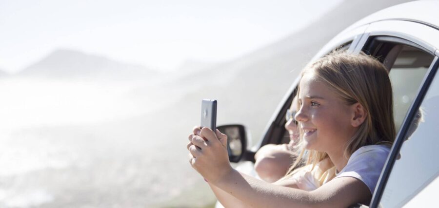 A young girl leans out of a car window, holding a smartphone and smiling, with a blurred outdoor landscape in the background.