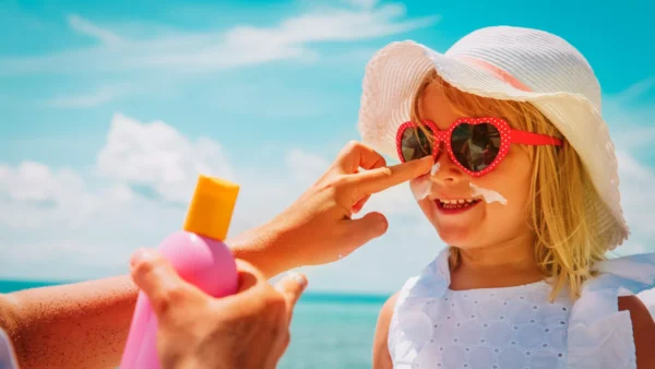 An adult applies sunscreen to a smiling child wearing a white hat and red heart-shaped sunglasses at the beach.