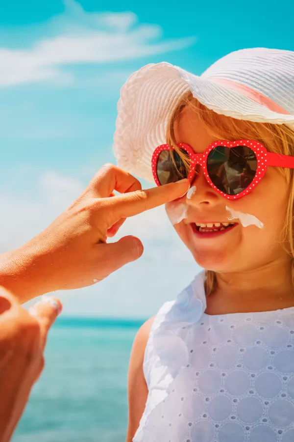 An adult applies sunscreen to a smiling child wearing a sunhat, heart-shaped sunglasses, and a white dress at the beach.