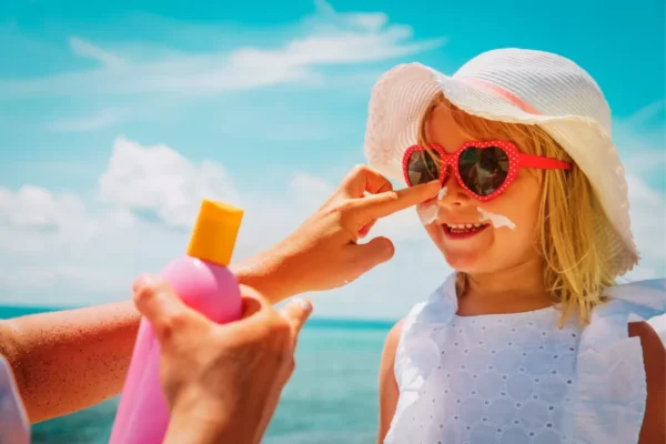 An adult applies sunscreen to a smiling child wearing a sun hat and heart-shaped sunglasses at the beach.