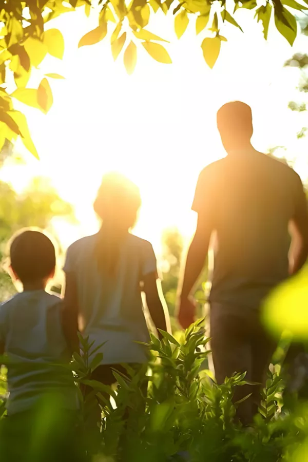 Three people, two children and one adult, walk through greenery towards bright sunlight, surrounded by leaves and plants.