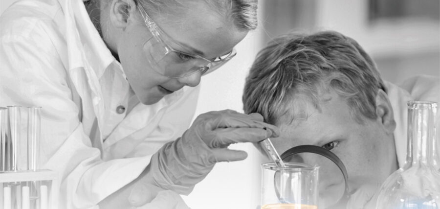 Two children in lab coats and safety goggles conduct a science experiment, using a pipette and a magnifying glass to examine a beaker.