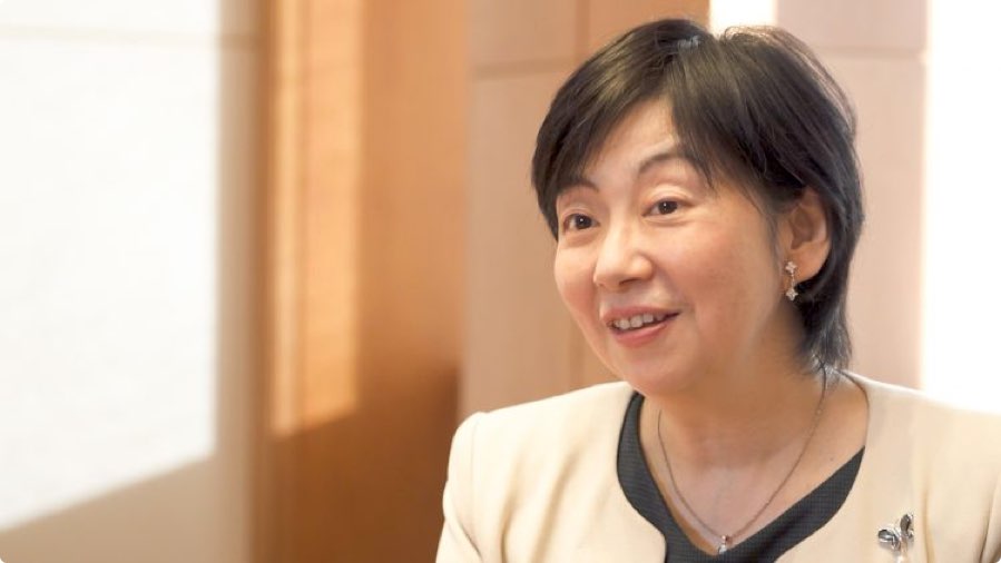 A woman with short dark hair, wearing a light-colored blazer and jewelry, is smiling while sitting indoors in a well-lit room.