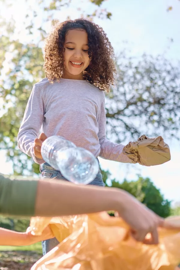 A child with curly hair holds recyclable items and helps place a plastic bottle into a large orange bag outdoors on a sunny day.