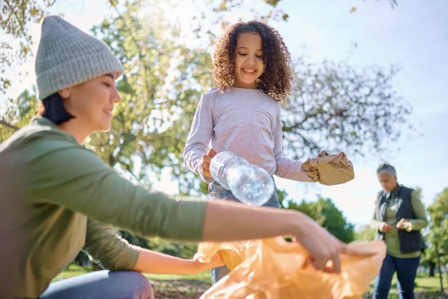 A child and an adult place litter into a bag outdoors, while another person picks up trash in the background on a sunny day.