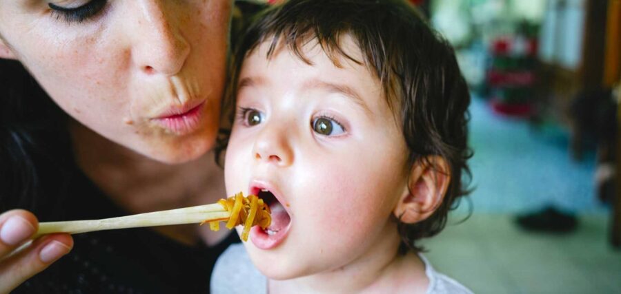 An adult holds pasta on a fork while blowing on it to cool it before feeding a young child with an open mouth.