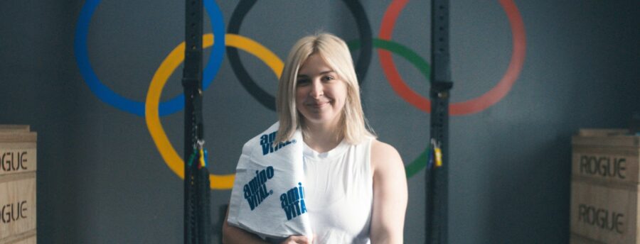 A woman with blonde hair holds a towel and stands in a gym with Olympic rings painted on the wall behind her and wooden boxes labeled "ROGUE" on each side.
