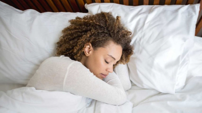 A person with curly hair is lying on their side, asleep in bed, covered with a white blanket and resting their head on a white pillow.