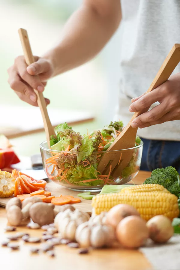A person uses wooden utensils to mix a bowl of salad with lettuce and carrots, surrounded by fresh vegetables on a wooden table.