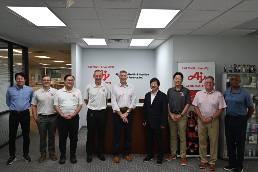 A group of nine men stand side by side in an office lobby in front of an Ajinomoto Health & Nutrition sign, posing for a group photo.
