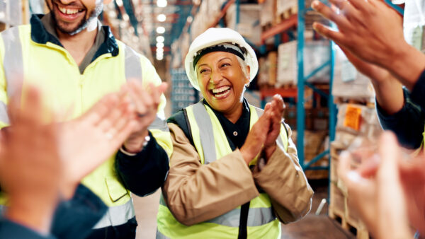 A smiling worker in a hard hat and high-visibility vest is applauded by colleagues in a warehouse setting.