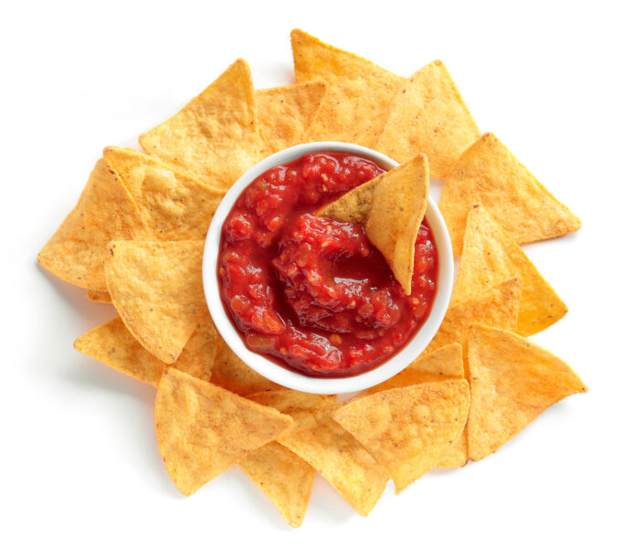 A bowl of tomato salsa surrounded by tortilla chips, with one chip dipped in the salsa, on a white background.