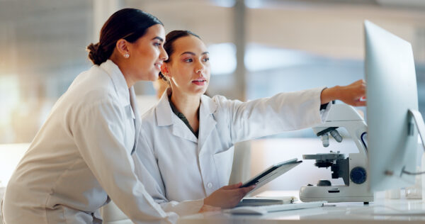 Two women in lab coats examine data on a computer monitor; one holds a tablet, and a microscope sits on the desk beside them.