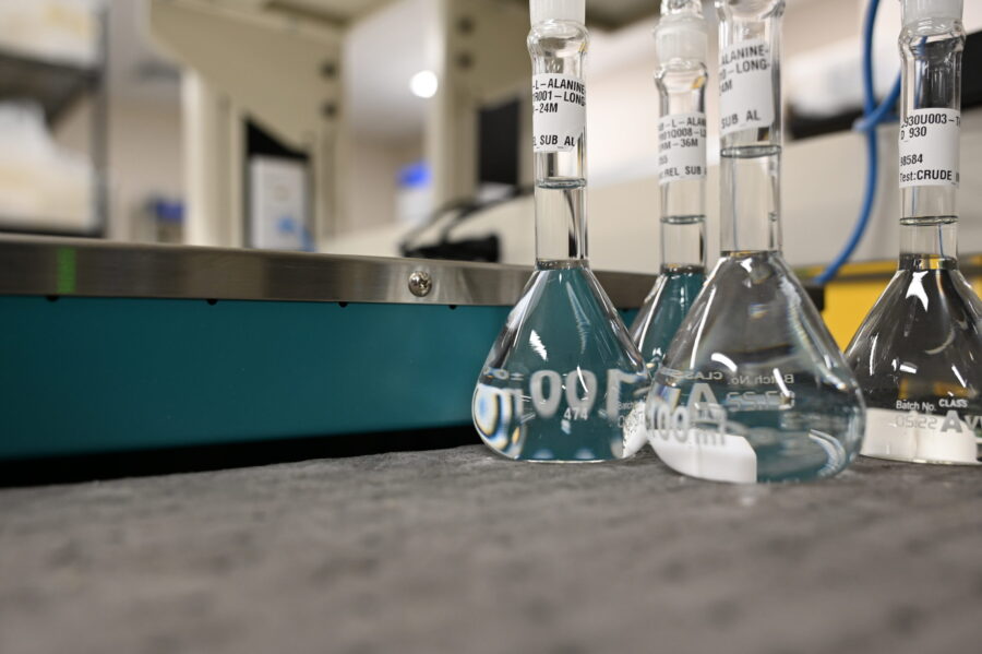 Three labeled glass Erlenmeyer flasks filled with clear liquid sit on a gray surface in a laboratory setting.