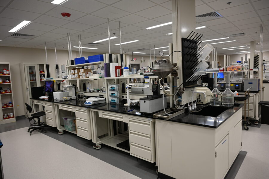 A modern laboratory with various scientific equipment, shelves with supplies, glassware, and storage cabinets along black countertops under bright fluorescent lighting.