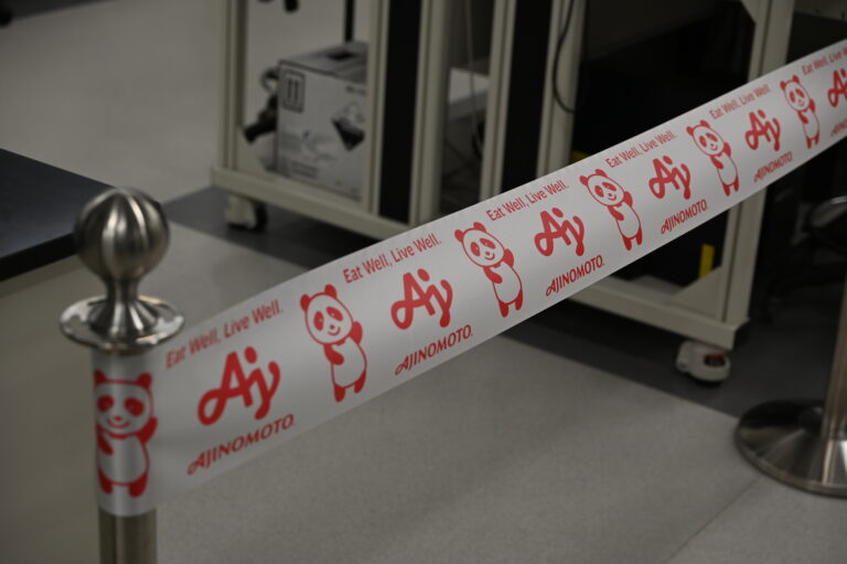 A white barrier tape with red Ajinomoto logos, panda illustrations, and the slogan "Eat Well, Live Well." blocks access in an indoor setting.