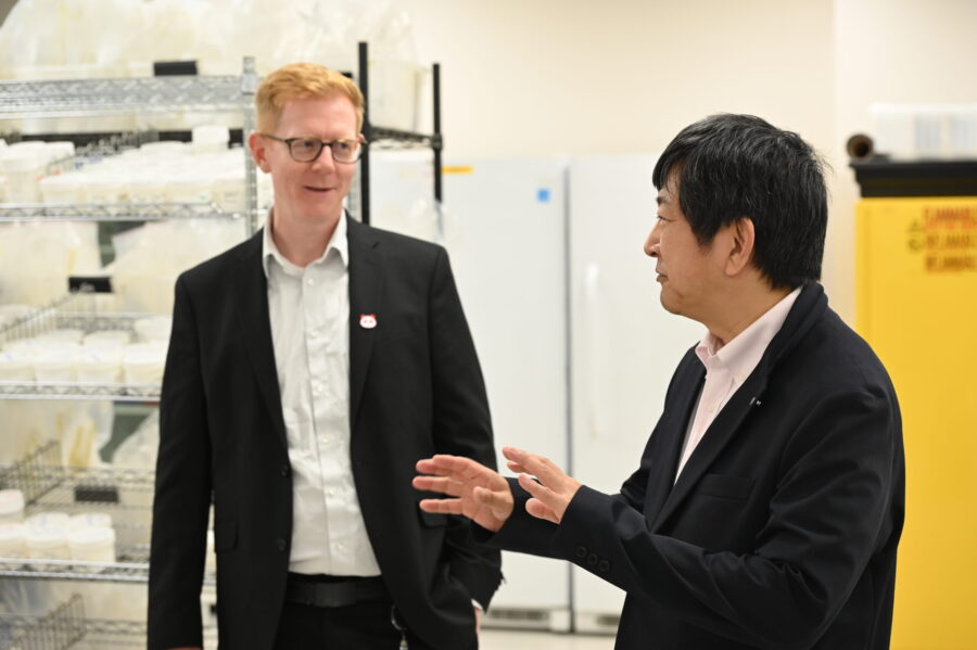 Two men in suits talk in a laboratory setting, with shelves of lab equipment and a yellow safety cabinet visible in the background.