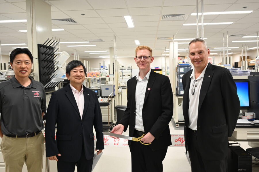 Four men stand in a laboratory, with one holding oversized scissors, suggesting a ribbon-cutting or ceremonial event. Laboratory equipment and computers are visible in the background.