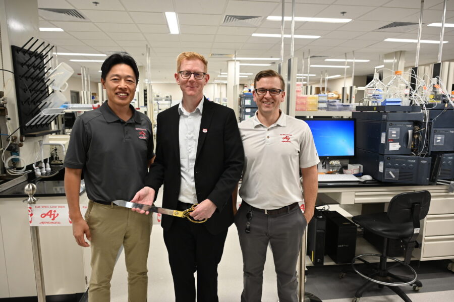 Three men stand together in a laboratory. One holds a ceremonial cake knife. Lab equipment and computers are visible in the background.