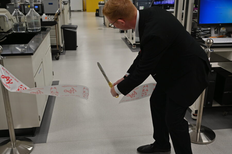 A person in a suit cuts a ceremonial ribbon with large scissors inside a laboratory setting.