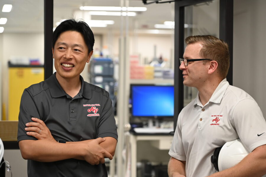 Two men in collared shirts stand and talk in an office or lab setting; one holds a white hard hat, and computers and equipment are visible in the background.