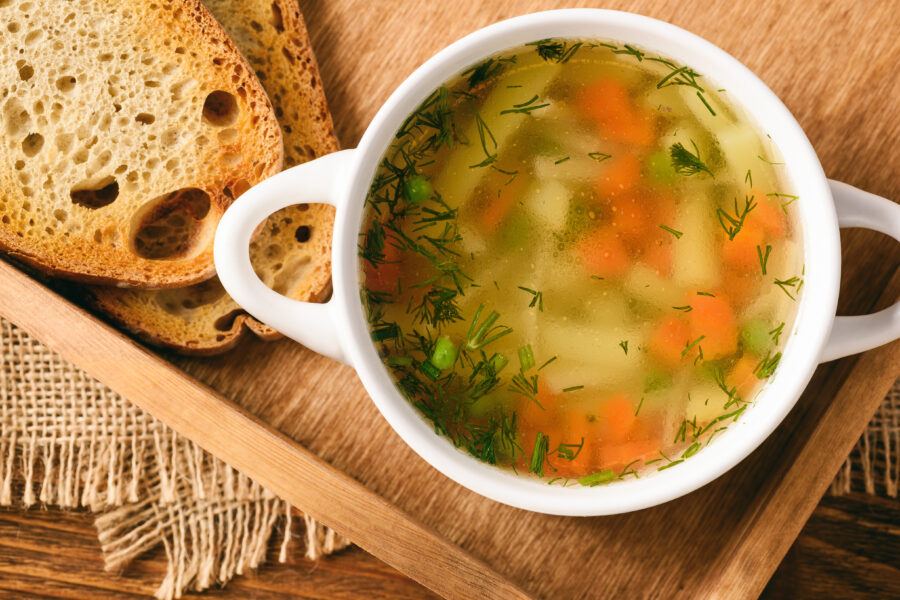 A bowl of vegetable soup with herbs, served on a wooden tray next to two slices of rustic bread.