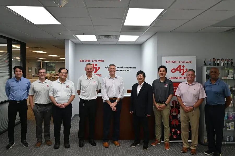 A group of nine men stand side by side in an office lobby in front of an Ajinomoto Health & Nutrition sign, posing for a group photo.