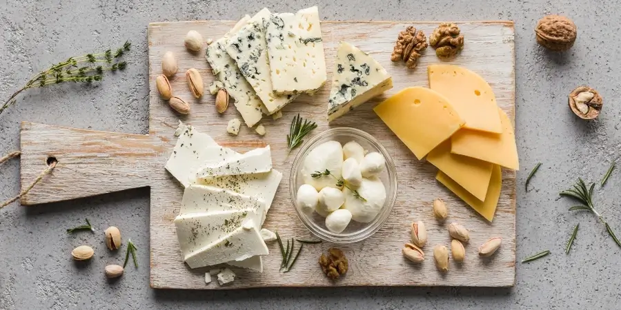 A wooden serving board with assorted cheeses, including blue cheese, feta, mozzarella balls, and sliced cheese, surrounded by nuts and sprigs of herbs.