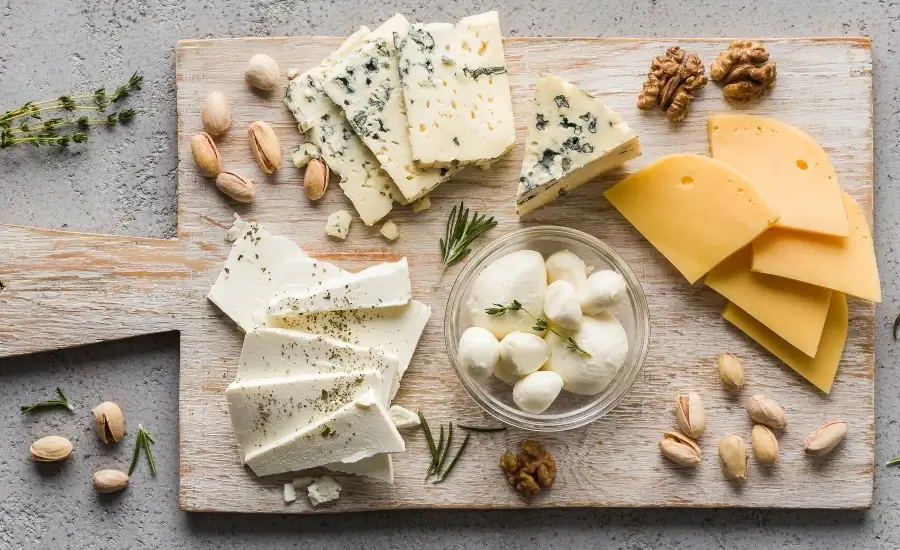 A wooden board with assorted cheeses, including blue cheese, mozzarella balls, feta, and gouda slices, surrounded by walnuts, pistachios, and herbs.