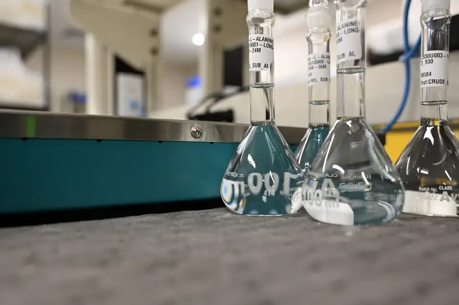 Three labeled glass Erlenmeyer flasks filled with clear liquid sit on a gray surface in a laboratory setting.