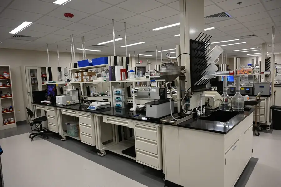 A modern laboratory with various scientific equipment, shelves with supplies, glassware, and storage cabinets along black countertops under bright fluorescent lighting.