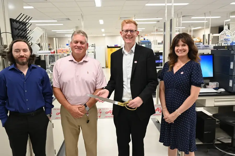 Four people stand in a laboratory; one person holds a ceremonial ribbon-cutting scissors.