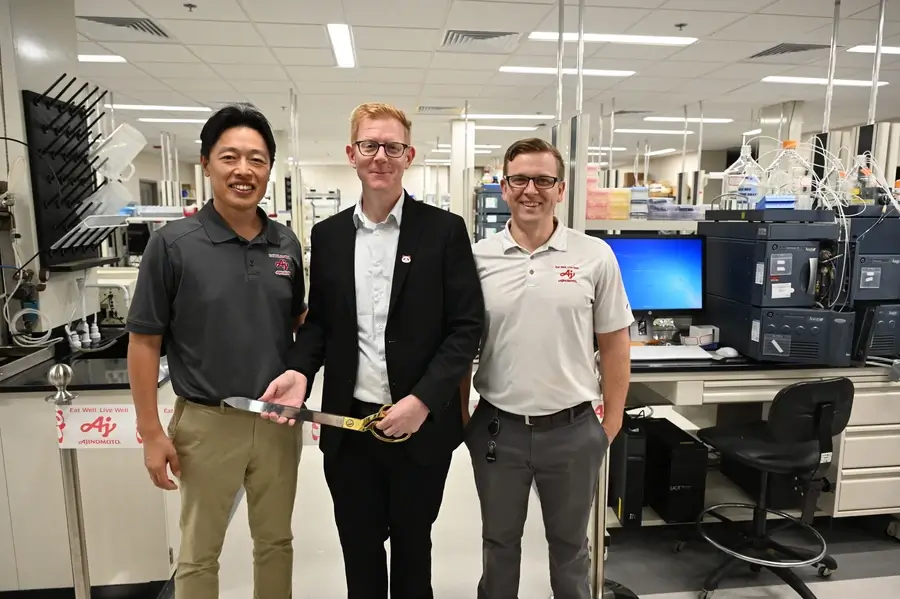Three men stand together in a laboratory. One holds a ceremonial cake knife. Lab equipment and computers are visible in the background.