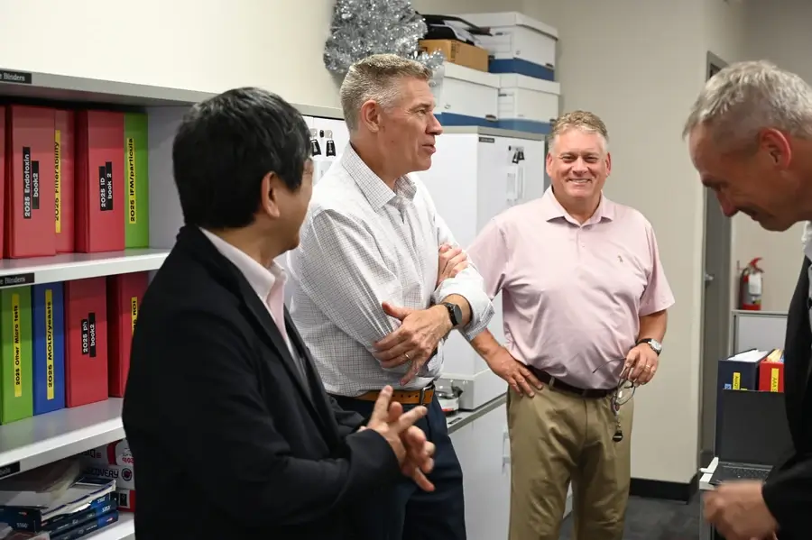 Four men stand and talk together in an office, surrounded by shelves with red and yellow files and office supplies.