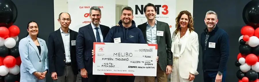 Seven people stand together, smiling, as one person holds a large check for $15,000 made out to MeliBio at an event with branded banners and balloons in the background.
