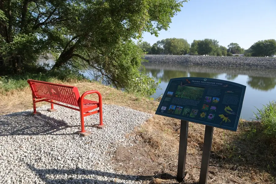 A red bench and informational sign about pollinators are positioned on gravel beside a calm river, with trees and greenery in the background.