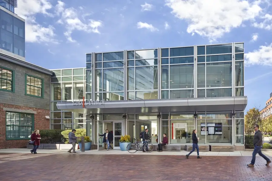 A modern glass building labeled "LabCentral" with people walking, a bicycle parked outside, and a partly cloudy sky in the background.