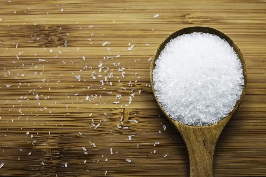 A wooden spoon filled with white, crystalline granules sits on a wooden surface, with some granules scattered around it.